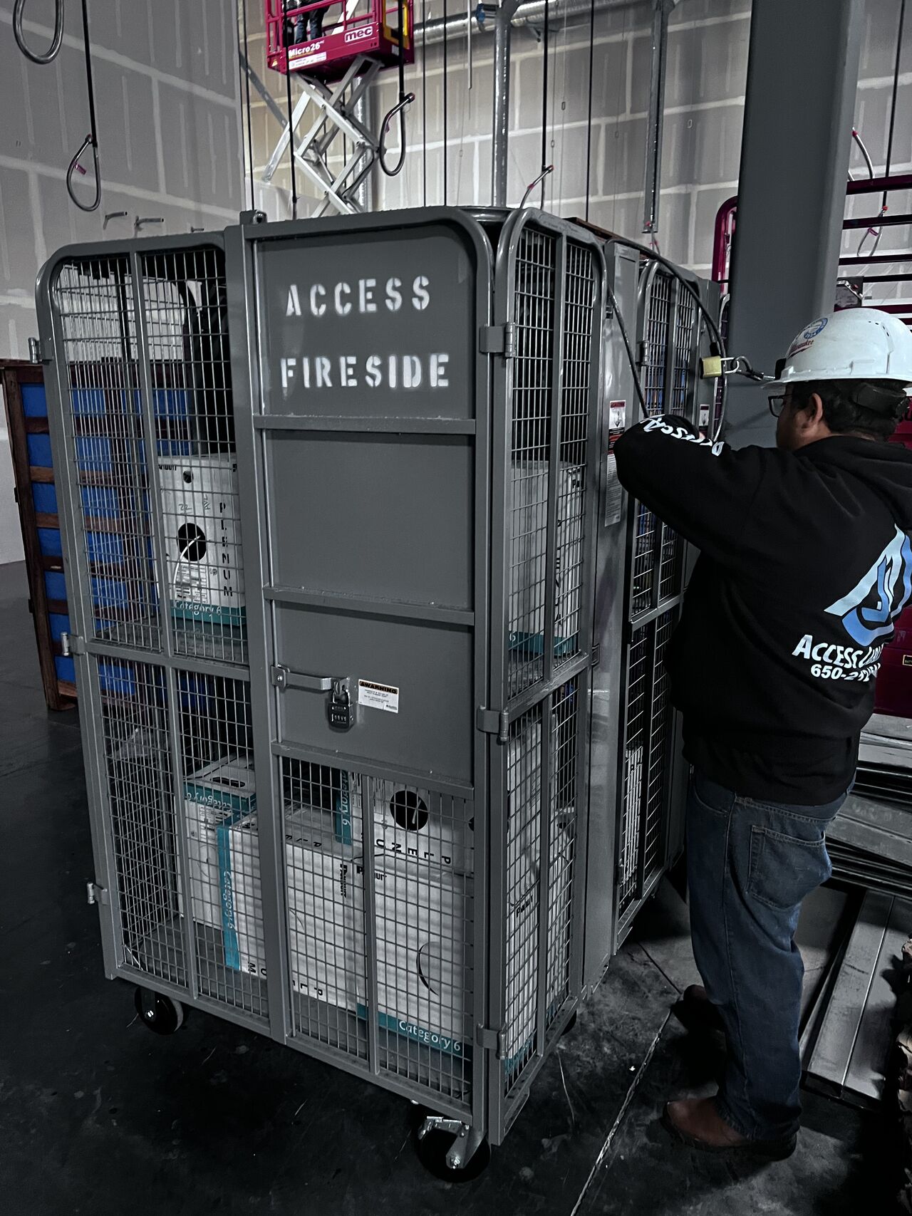 Padlocked Access Fireside job-site security cage at the Amazon Rancho Cordova fulfillment facility loaded with boxes of Panduit Category 6 plenum cable, with an Access Cabling technician in branded 650 hoodie securing the door.