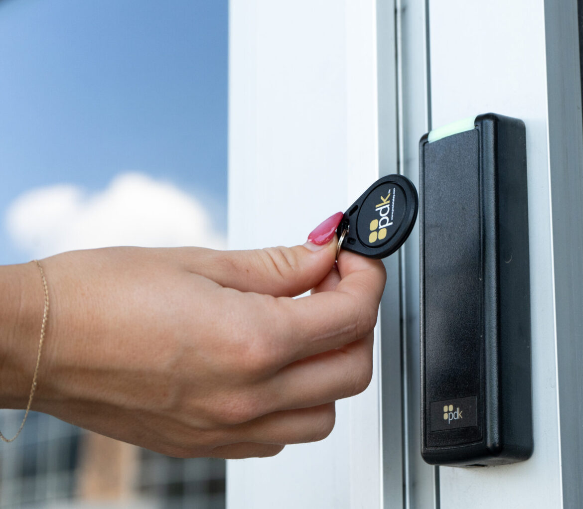 Person presenting a PDK ProdataKey fob to a PDK wall-mounted reader at a commercial glass entry door.