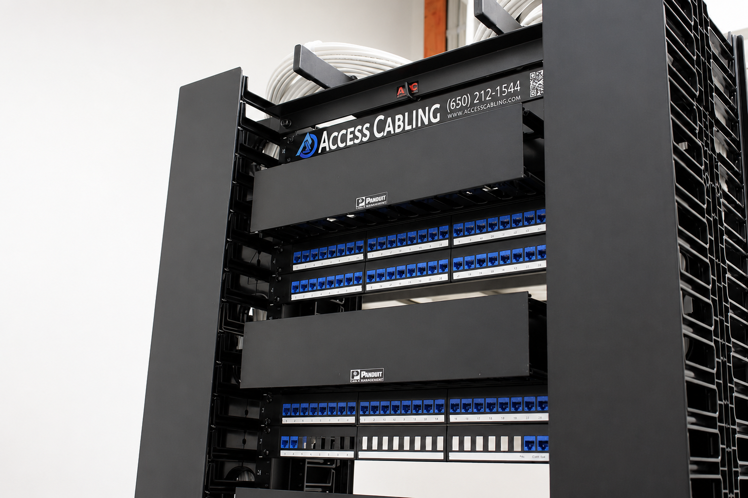 Access Cabling-branded Panduit enterprise rack with stacked blue Category 6 patch panels, horizontal cable managers and white station cables coiled above the cabinet — installed during a data center rack buildout.