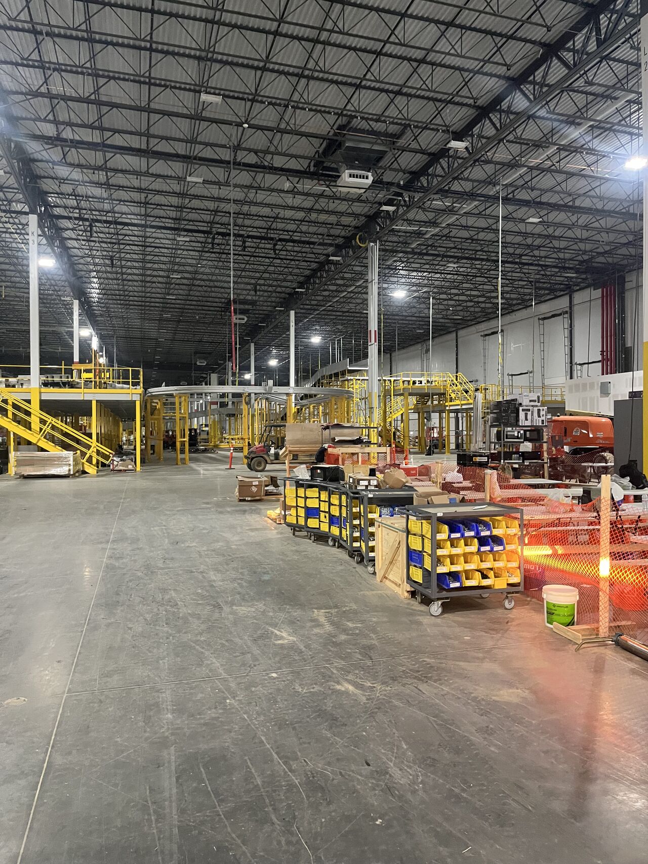 Interior of the Amazon Rancho Cordova fulfillment facility during construction, showing the yellow structural mezzanine, exposed bar-joist roof deck, curved conveyor take-away and staged material carts loaded with hardware bins — the operations floor where Access Cabling ran the tray, J-hook pathways and Cat6A drops.